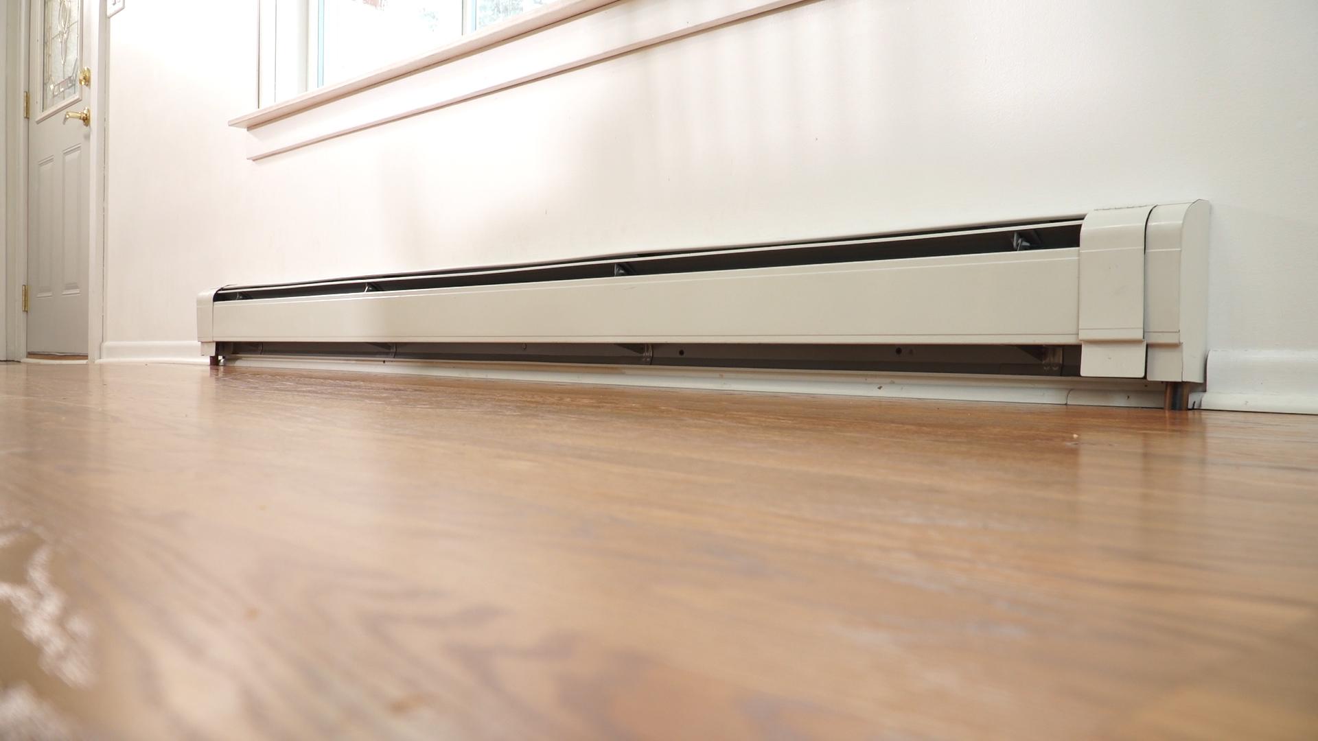 Long white baseboard heater installed along a wall with hardwood flooring, located below a window